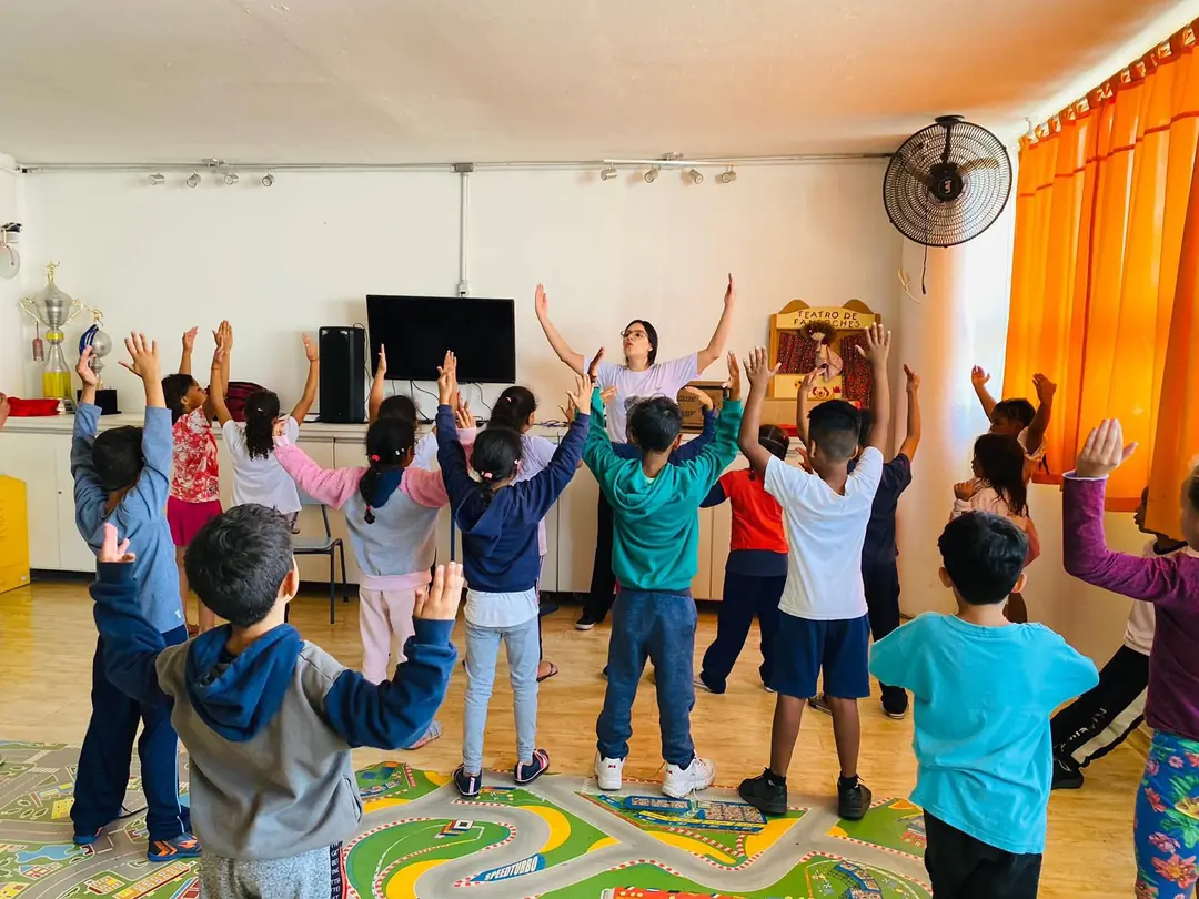 Prof. Sabrina conduzindo uma turma de crianças com os braços erguidos durante atividade de musicalização